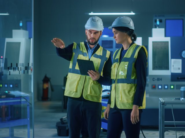 Modern,Factory:,Female,And,Male,Engineers,Wearing,Safety,Jackets,,Hardhats